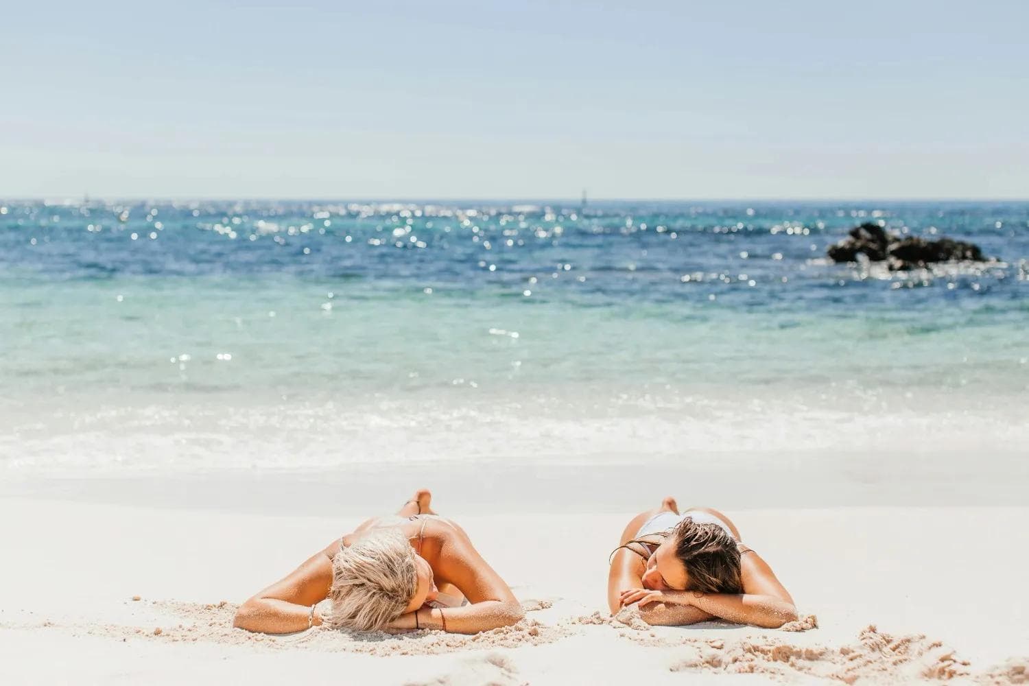 Two women sunbathing on a white sand tropical beach with clear blue ocean. Best beach holidays, summer escape, Dubai tour package options.